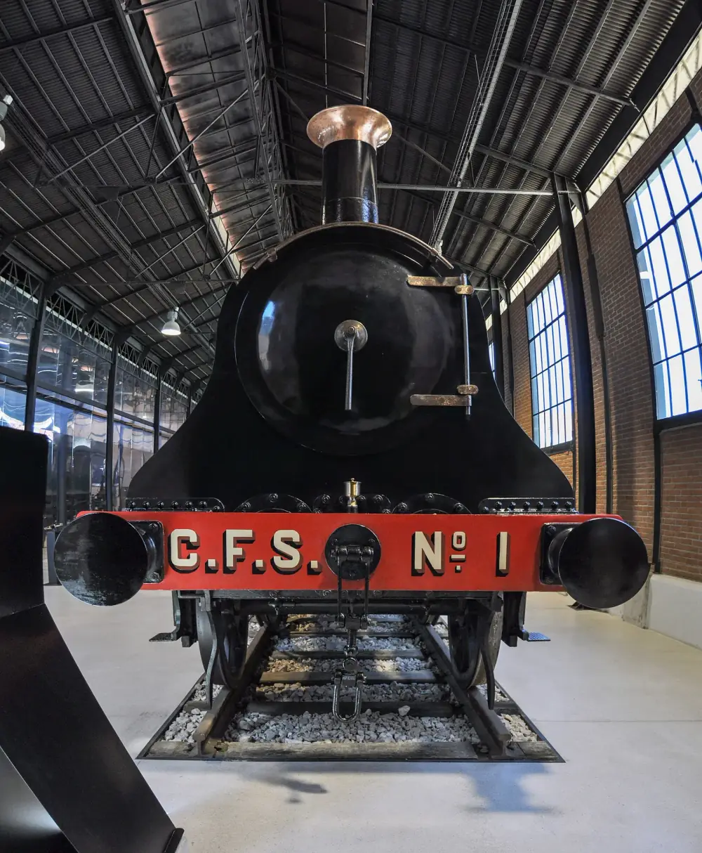 Front view of a black steam locomotive with a tall chimney and a red buffer beam lettered C.F.S. No. 1, standing on a short length of track inside a long hall with a steel roof and tall windows.