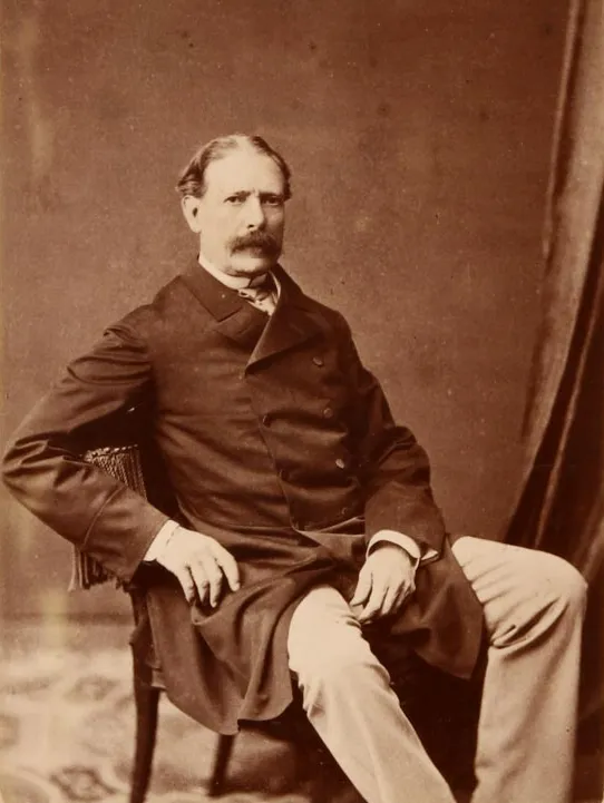 Sepia studio photograph of a seated middle-aged man with a thick moustache, in a dark frock coat and light trousers, one arm resting on the back of a fringed chair.