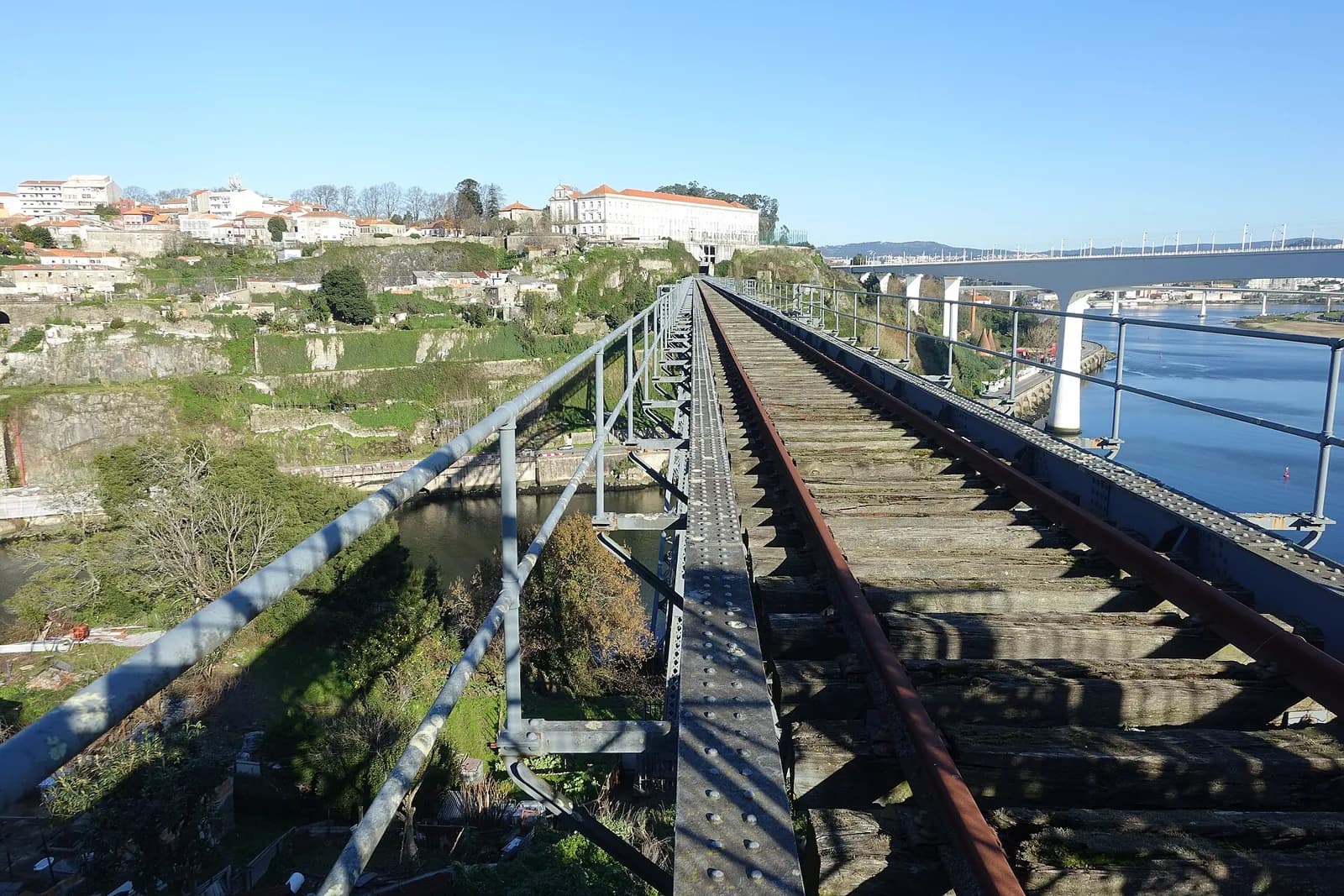 A single rusty railway track runs between metal railings across a high iron bridge. Below on the left are a river bank, a quarry face and houses on a hillside; on the right a modern concrete bridge on white piers runs alongside above the river, under a clear sky.