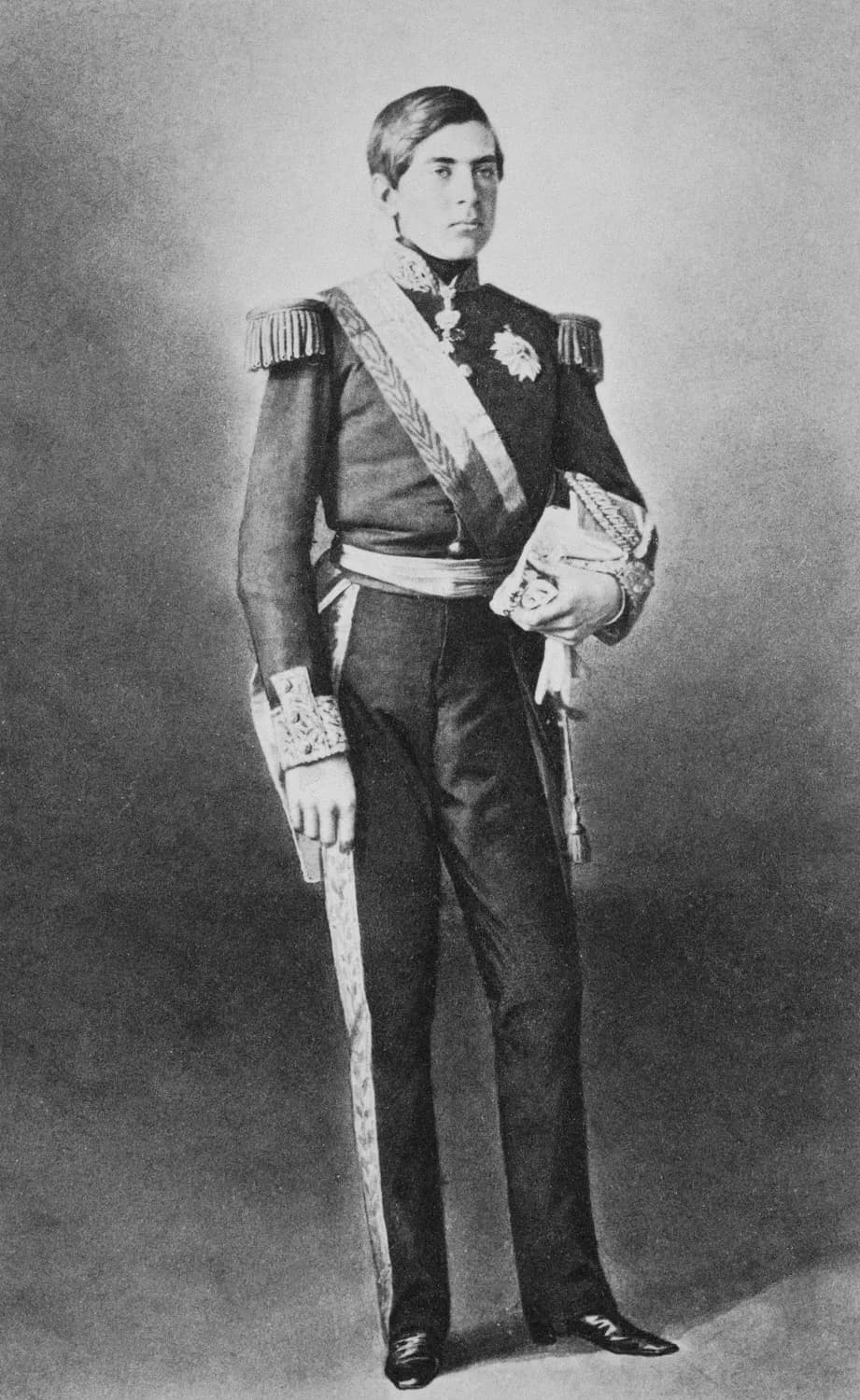 Full-length photograph of a slim young man standing in a dark military uniform with fringed epaulettes, embroidered cuffs, a sash and a breast star, holding a plumed hat under one arm.