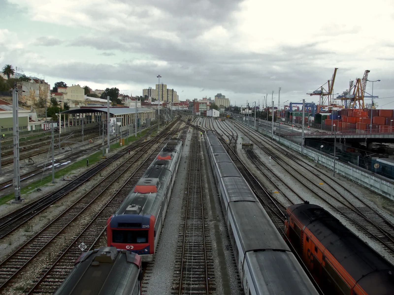Seen from above, many tracks fan out under a cloudy sky. Suburban trains in white, red and blue and an orange train stand in the sidings, with port cranes and stacked containers on the right.