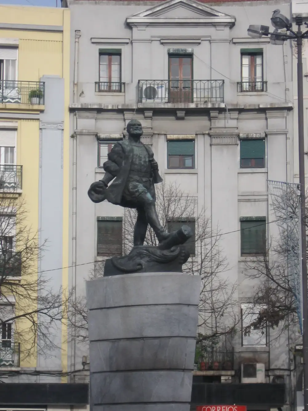 A dark bronze statue of a bearded man in sixteenth-century dress striding forward with a hat in one hand, on a tall grey stone pedestal in front of a pale apartment building.