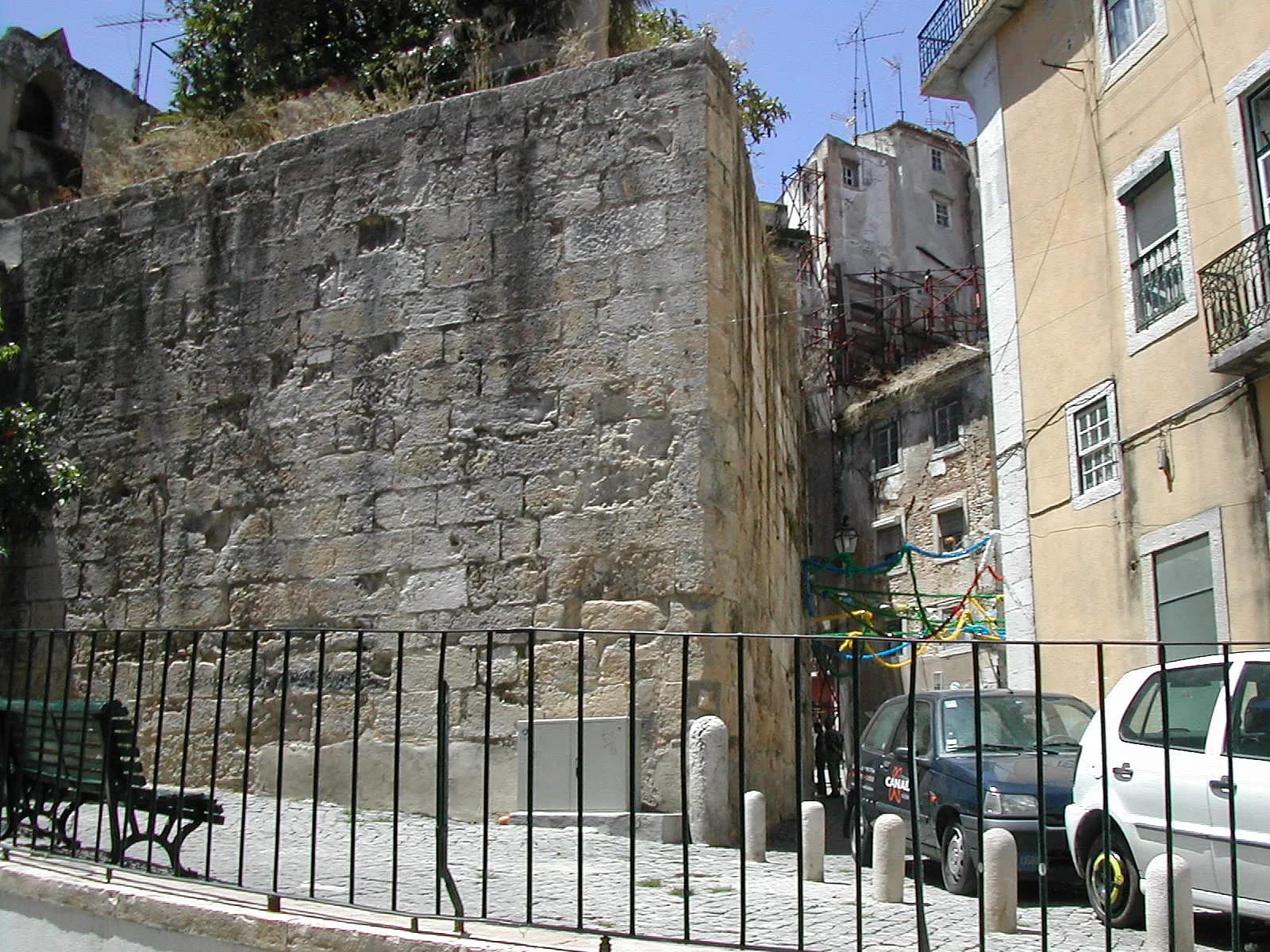 A massive square tower of rough, weathered limestone blocks rises from a cobbled square behind an iron railing. Narrow old apartment buildings and parked cars crowd beside it.