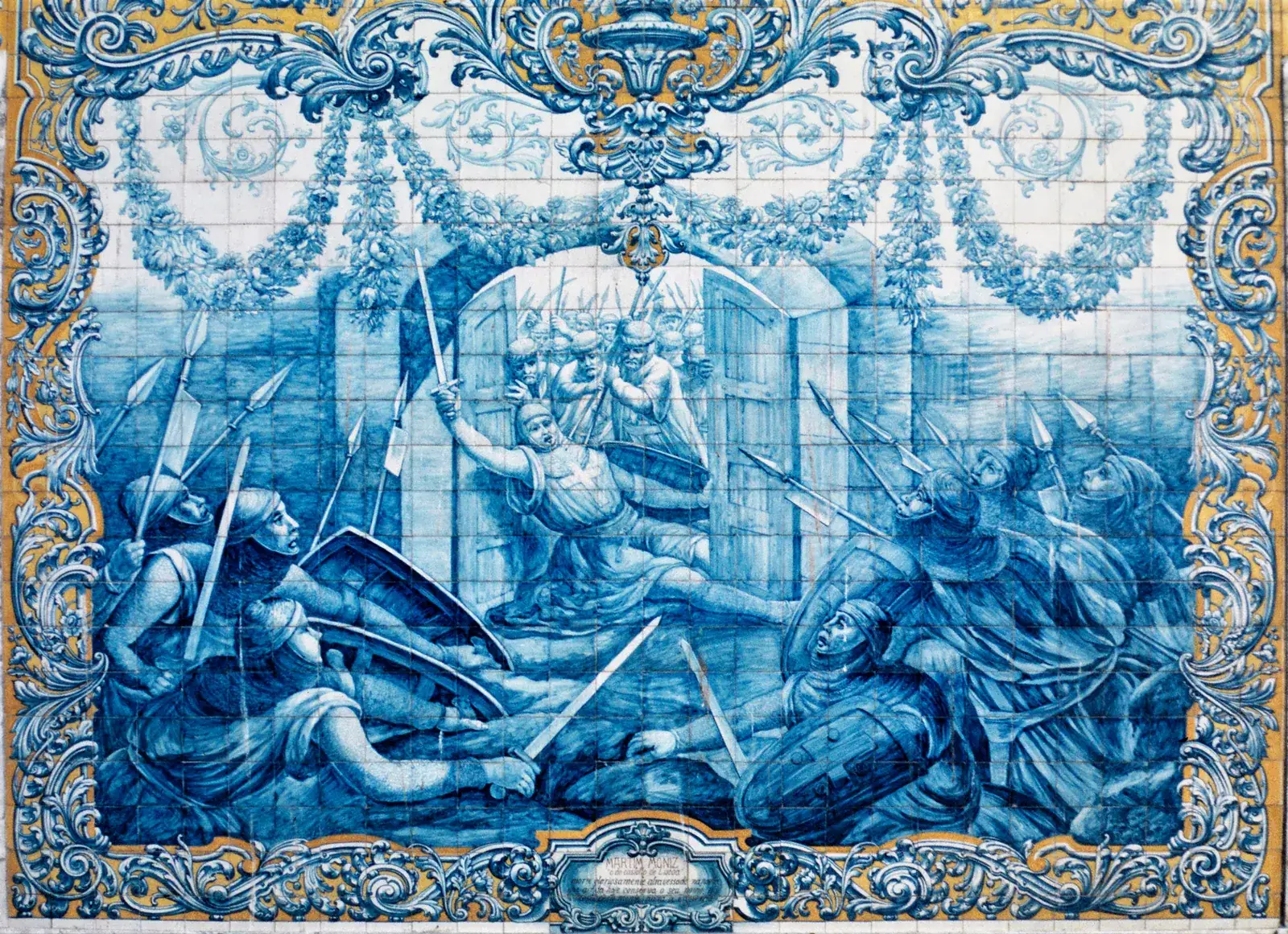 A blue-and-white tile panel inside an ornate frame of scrolls and garlands picked out in yellow. A knight with a cross on his tunic falls backwards between the two wooden leaves of a gate, sword raised, while defenders with spears push from inside and armed men with shields and spears crowd in from both sides.