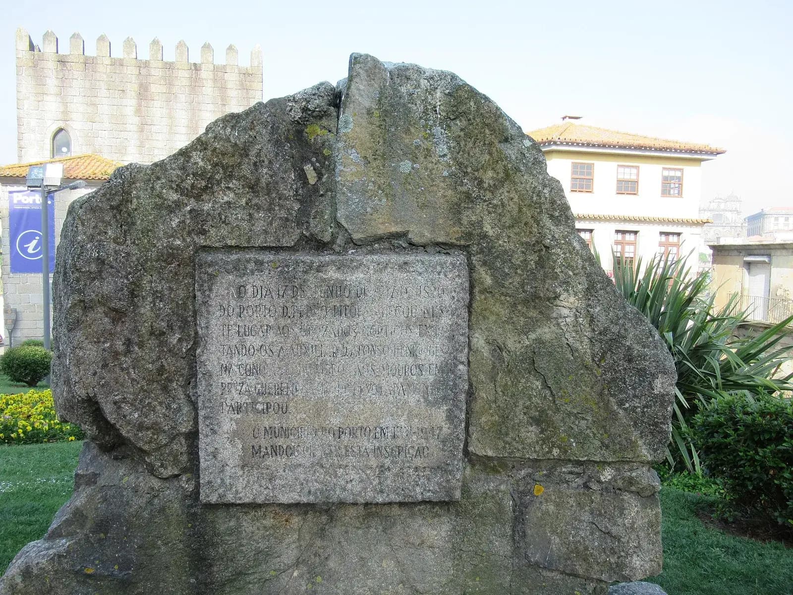 A large rough granite boulder set in a small garden, with a weathered rectangular stone plaque carved with capital letters. A crenellated stone tower and a white house with a tiled roof stand behind it.