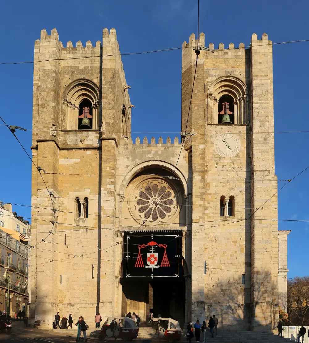 The west front of a stone cathedral with two square crenellated bell towers framing a large rose window over a deep arched portal, under a clear blue sky. Tram wires cross the facade and people walk past at street level.