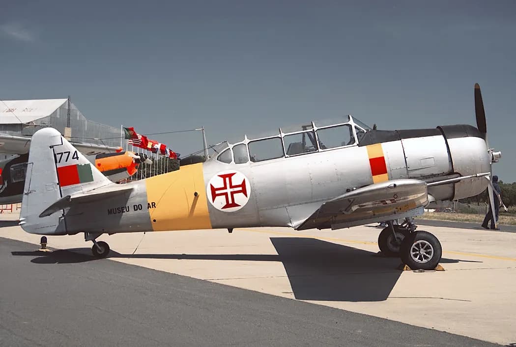 A silver single-engine propeller trainer with a yellow band round the fuselage, the red Cross of Christ roundel, the words Museu do Ar and the tail number 1774, partly cut off, parked on an airfield apron with flags behind it.