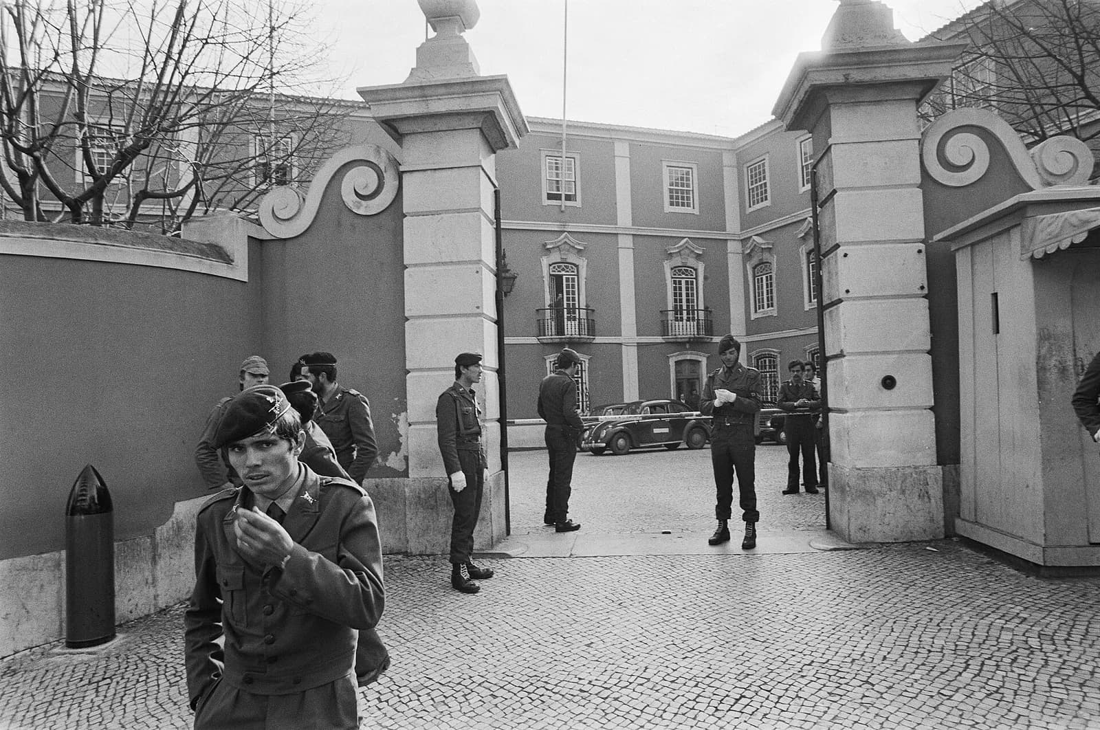 Black-and-white photograph of young soldiers in berets and field uniforms standing around an ornate stone gateway with scrolled pillars. Behind them are a cobbled courtyard, parked cars and a three-storey building with balconied windows. A soldier in the foreground holds a hand to his mouth.