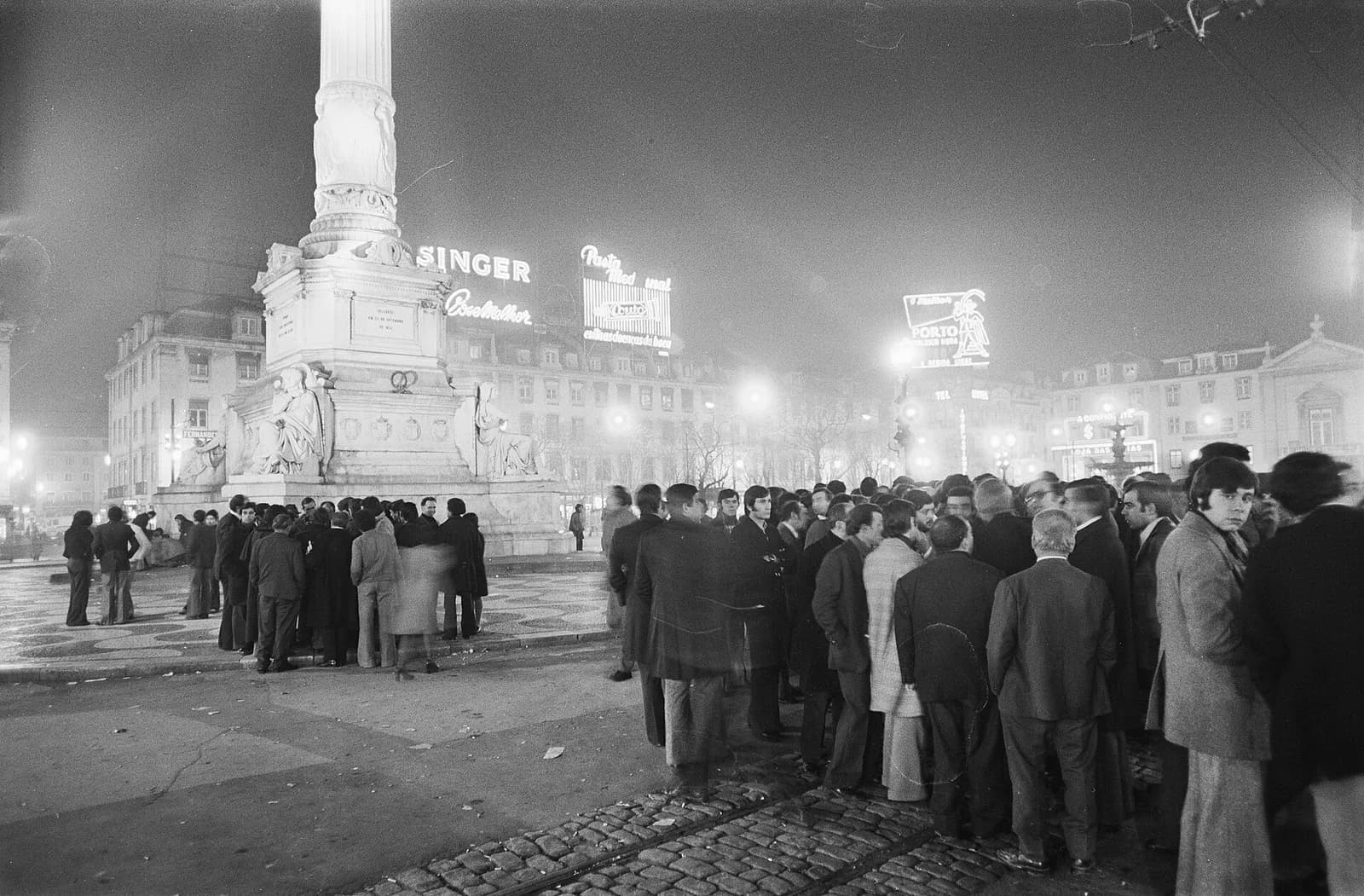 Night photograph of a crowd of men in dark coats standing on a cobbled square, with a tall floodlit column and lit-up advertising signs, one reading Singer, on the buildings behind.