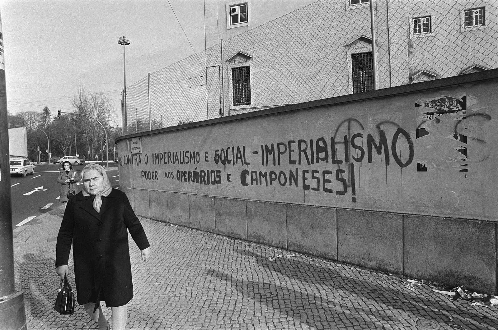 A woman in a dark coat and headscarf, handbag on her arm, walks along a pavement past a long low wall painted with large slogans; a wire fence and a building rise behind the wall.