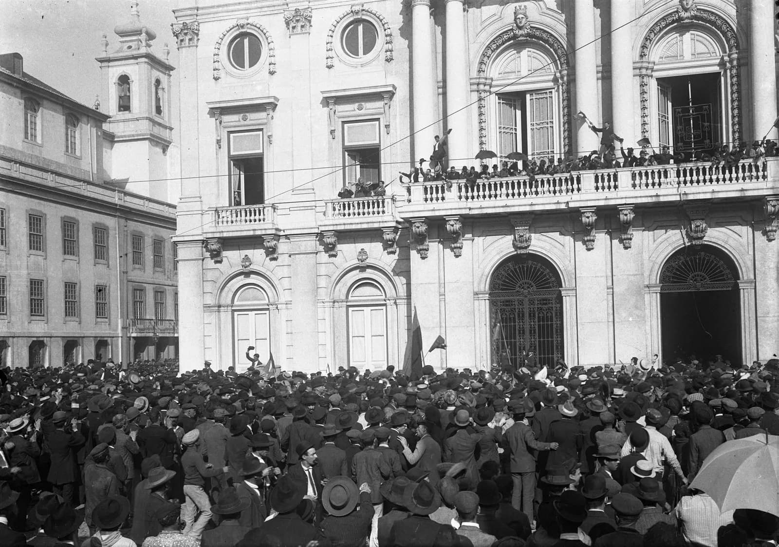 A dense crowd of men in hats fills a square in front of a large neoclassical building. Men pack the long stone balcony above, one waving his hat, and two flags rise from the crowd.
