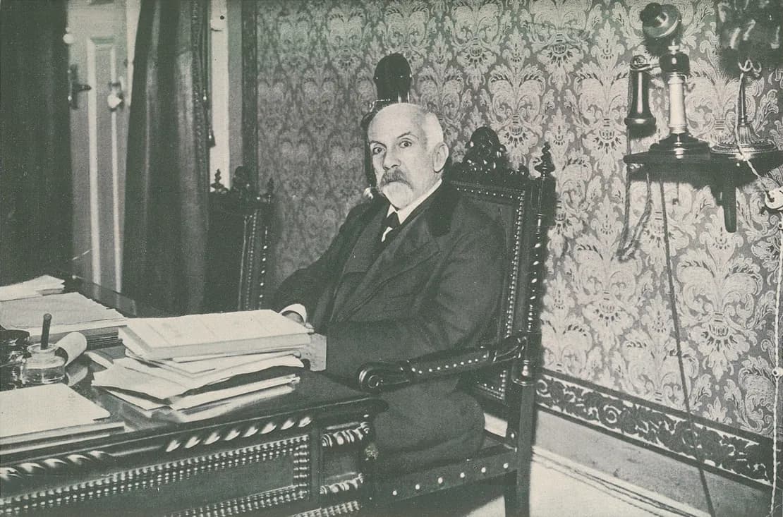 An elderly man with white hair and a grey moustache sits in a carved high-backed chair at a desk stacked with papers, in a room with patterned wallpaper and a wall telephone.