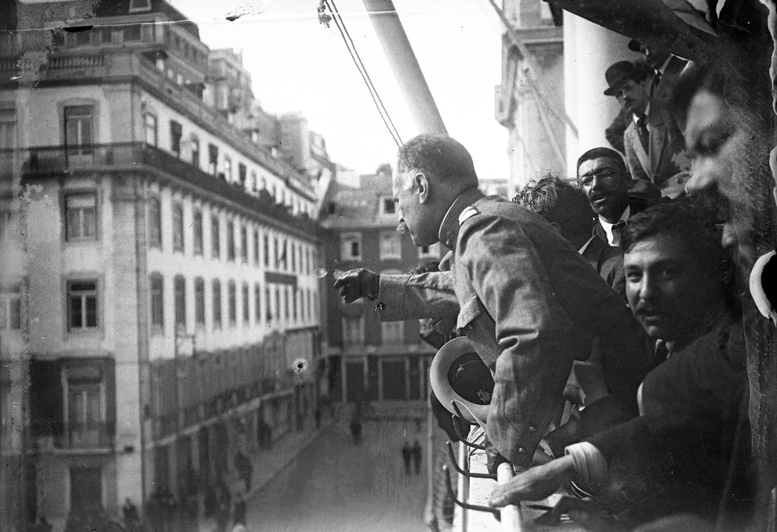 A grey-haired officer in uniform leans over a balcony railing and points down at a street lined with tall buildings, surrounded by men in civilian clothes pressing in beside him.