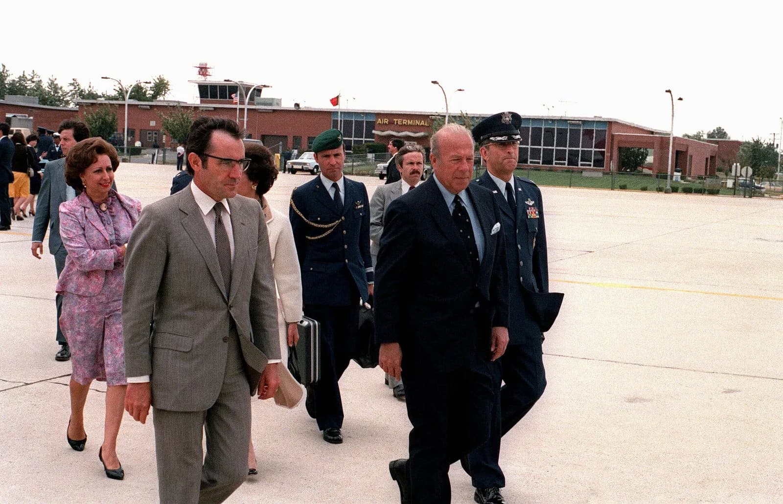 Colour photograph of a group walking across an airfield apron. In front on the left is a slim man in a grey suit and glasses, with women in pink and white behind him; on the right walk an older man in a dark suit and a uniformed officer. A building marked Air Terminal is behind them.