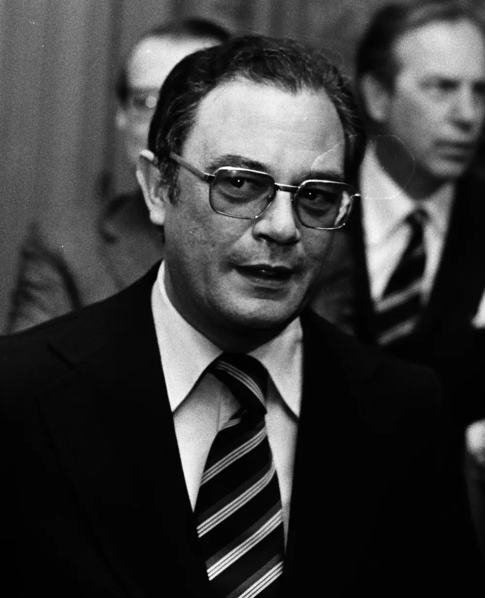 Black-and-white photograph of a man in his forties with dark-rimmed glasses, a dark suit and a striped tie, speaking. Another man in a suit stands behind him to the right.