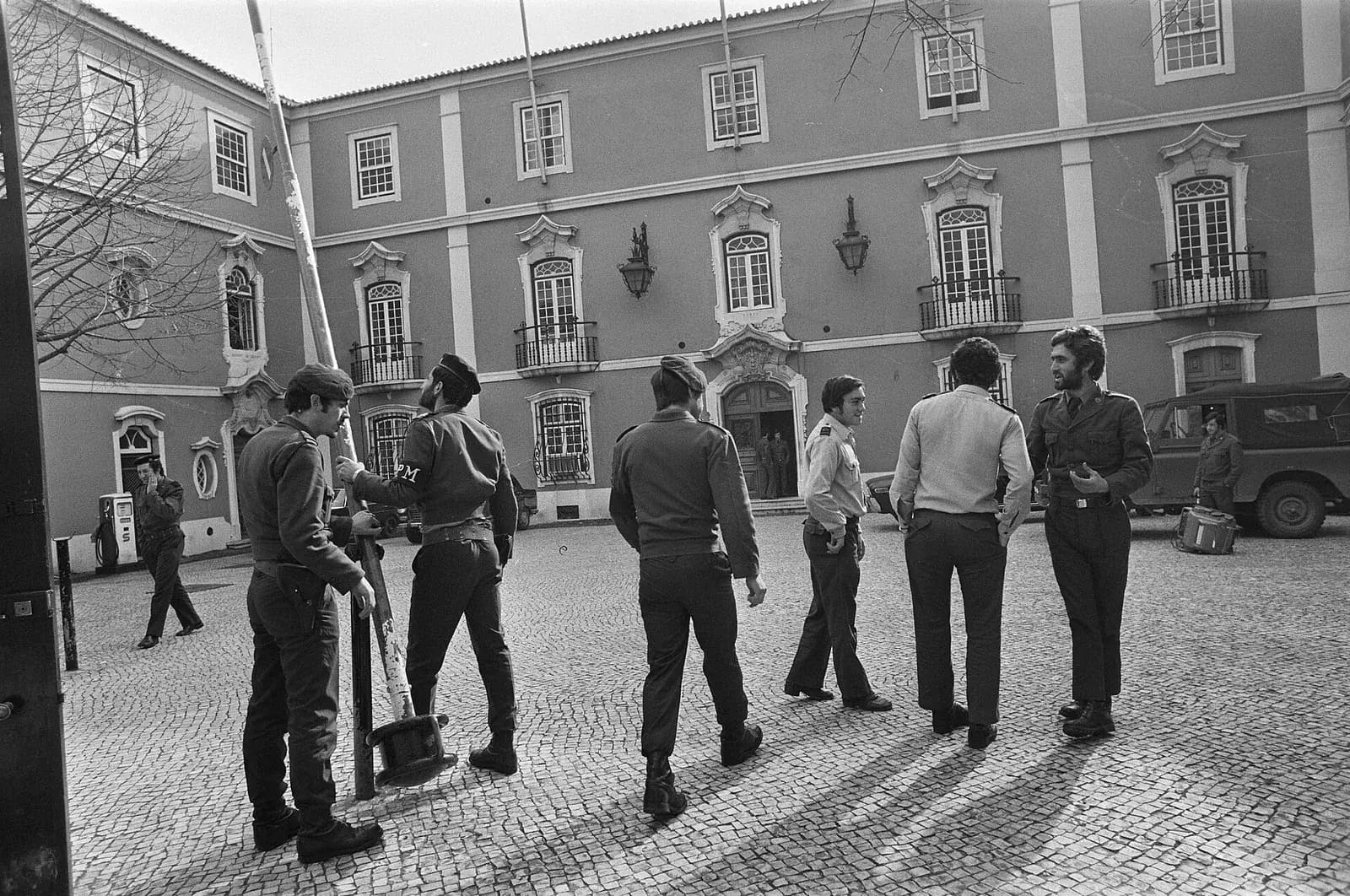 Black-and-white photograph of a cobbled courtyard in front of a three-storey building with ornate window frames and iron balconies. A group of soldiers in berets and fatigues stands talking near a barrier pole; one wears an armband marked PM. Two men in shirtsleeves and a jeep are on the right.