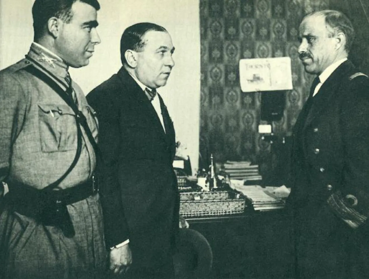 Black-and-white photograph of three men talking in an office: on the left a young officer in a light uniform with a shoulder strap, in the middle a man in a dark suit, and on the right an older man with a moustache in a dark naval uniform.