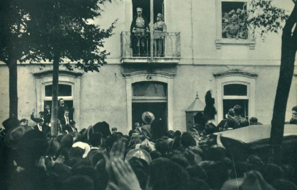 Black-and-white photograph of a crowd in a street below a building; two uniformed men stand on a first-floor balcony and others watch from a window.