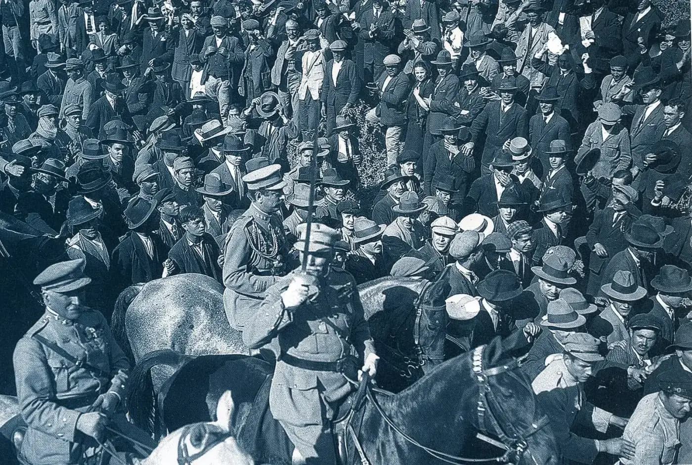 Black-and-white photograph of uniformed officers on horseback moving through a dense crowd of men in hats and caps; one rider holds a sword upright.
