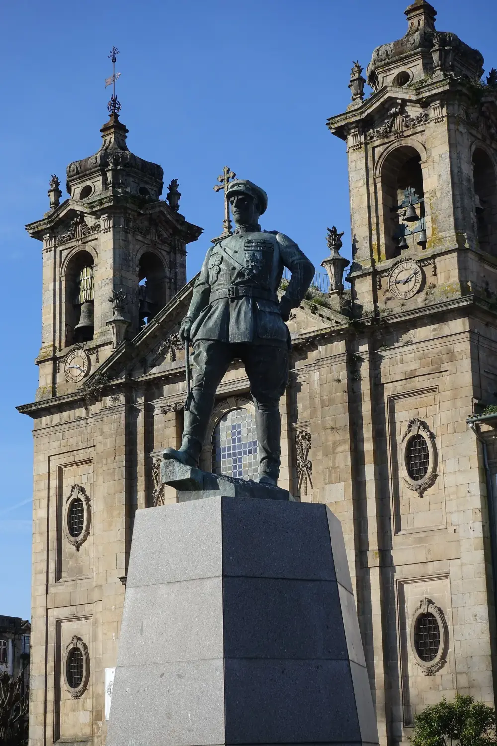 Bronze statue of a soldier in a peaked cap and field uniform on a tall grey stone pedestal, with a granite Baroque church facade and twin bell towers behind, under a blue sky.
