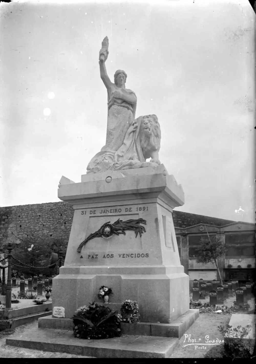 A stone monument in a cemetery: a woman holding up a torch stands beside a lion on a tall pedestal inscribed 31 de Janeiro de 1891 and A paz aos vencidos, with wreaths at its foot.