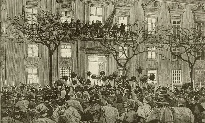 An engraving of a dense crowd raising hats and canes in front of a large building with bare trees. On a long first-floor balcony a row of men stands beside a furled flag.