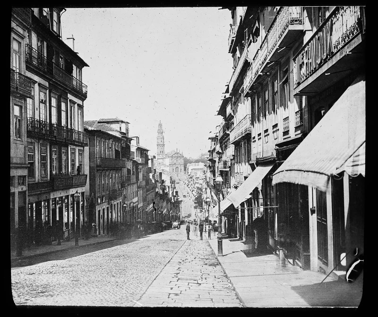 A black and white photograph of a narrow cobbled street sloping downhill between tall buildings with iron balconies and shop awnings. A few men stand in the roadway, and at the far end a tall baroque church tower rises above the rooftops.