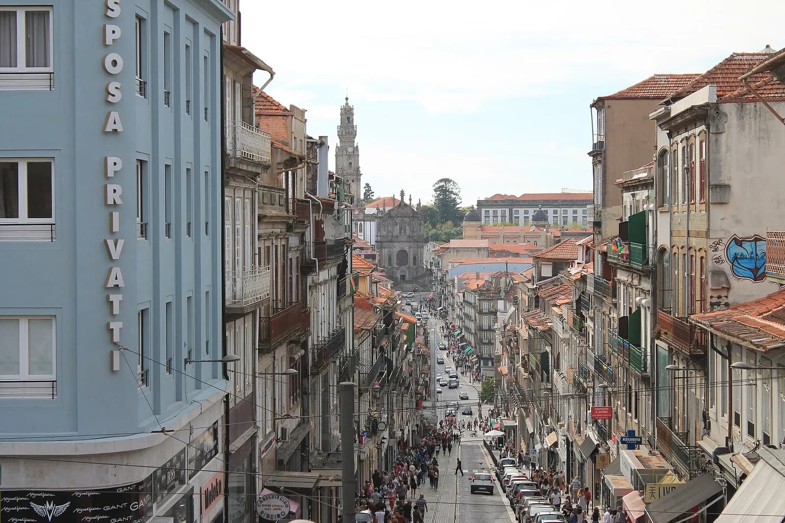 A busy, steep shopping street seen from above, lined with tall old buildings and crowded with pedestrians, with a baroque church tower on the skyline at the far end.