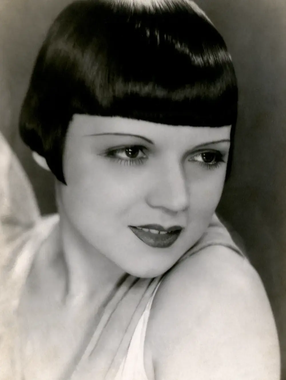 Studio portrait of a young woman with a glossy black bob and a straight fringe, looking over her bare shoulder with a slight smile.
