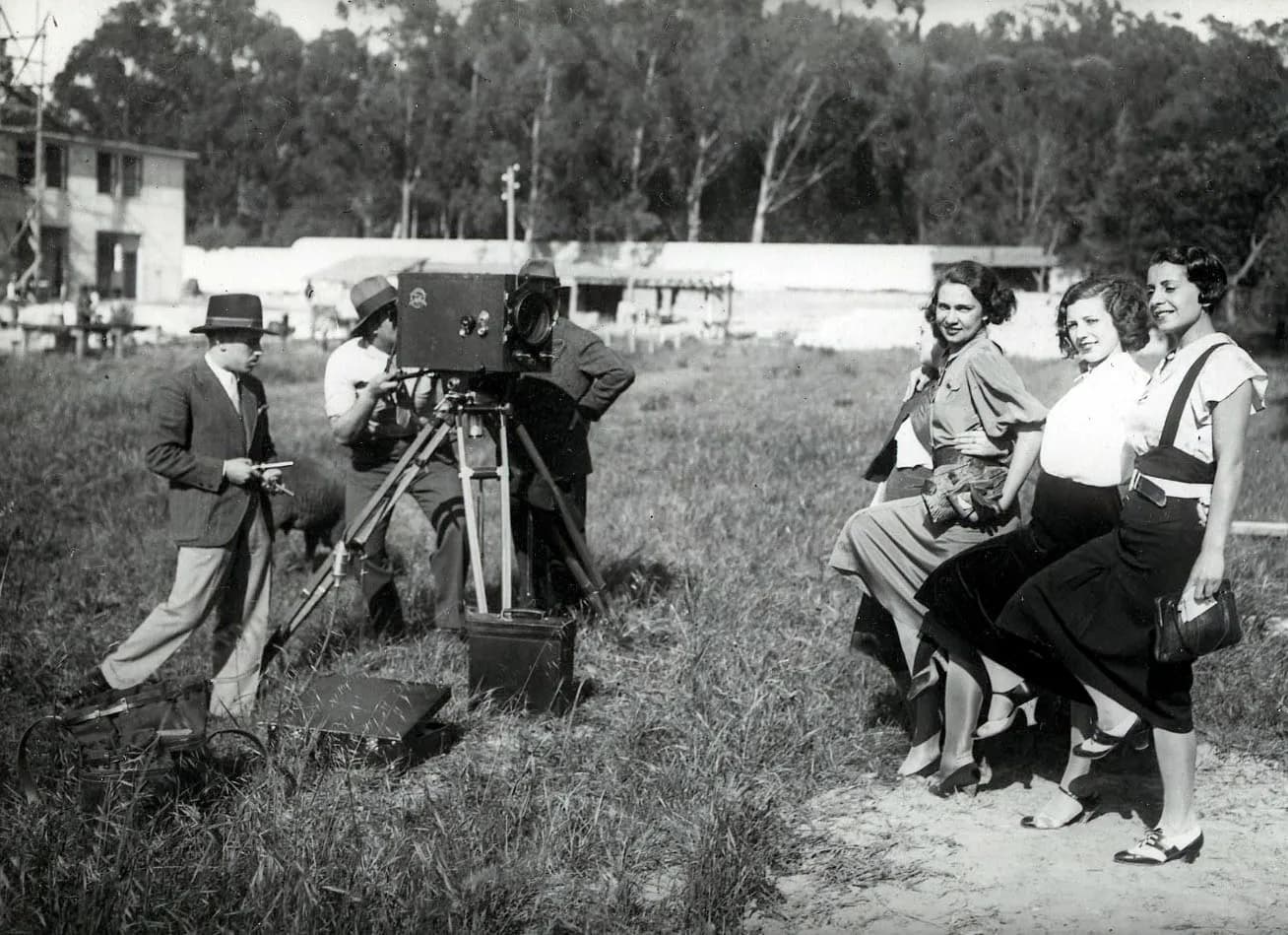 In a grassy field four young women in blouses and skirts, one almost hidden behind the others, stand arm in arm and kick one leg forward for a box film camera on a wooden tripod. Two men in hats work the camera, a man in a suit and trilby stands beside it holding a small object, and low white buildings and tall trees fill the background.