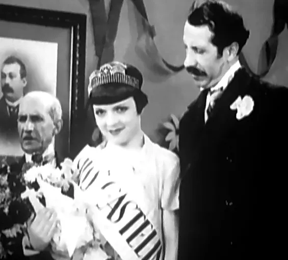 A young woman with a dark bob, a sparkling tiara and a sash reading CASTEL across her chest holds a bouquet. A moustached man in a dark suit with a white flower in his buttonhole stands at her side, an older white-haired man stands behind her to the left, and a framed portrait of a moustached man hangs on the wall.