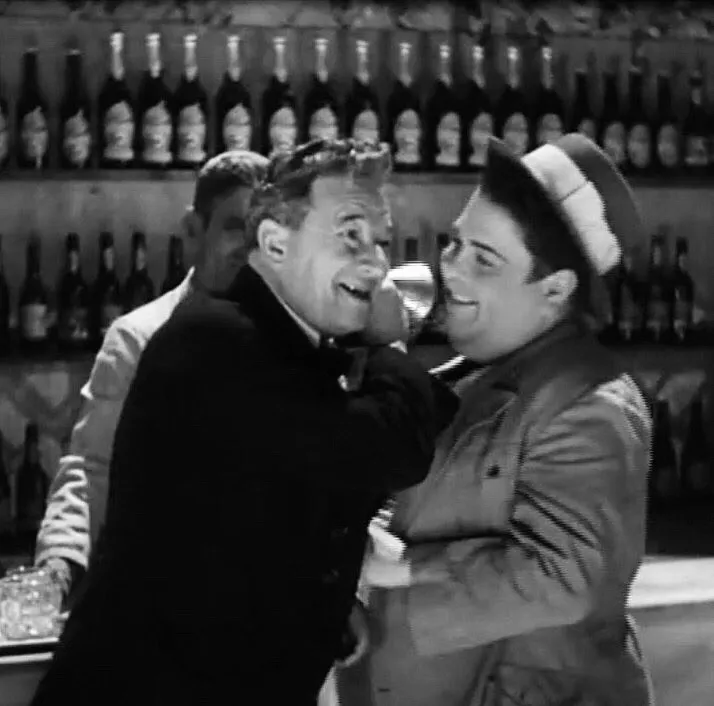 Two men laugh at a bar in front of shelves of bottles. The older man on the left, in a dark coat, cups a hand to his ear; the younger, plumper man on the right wears a straw boater and leans in close to him.