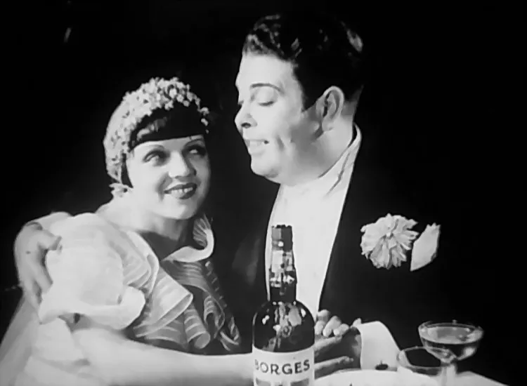 A plump groom in a tailcoat, with a large flower in his lapel, has his arm around a smiling bride in a flowered cap. On the table in front of them stand a wine bottle labelled BORGES and champagne glasses.