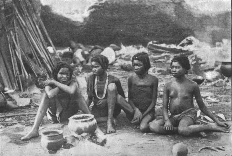 Black and white photograph of four young African women sitting on bare ground in front of a thatched shelter, with clay pots beside them.