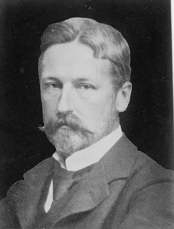 Black and white head-and-shoulders portrait of a man with swept-back hair, a moustache and a pointed beard, wearing a dark jacket and high white collar.