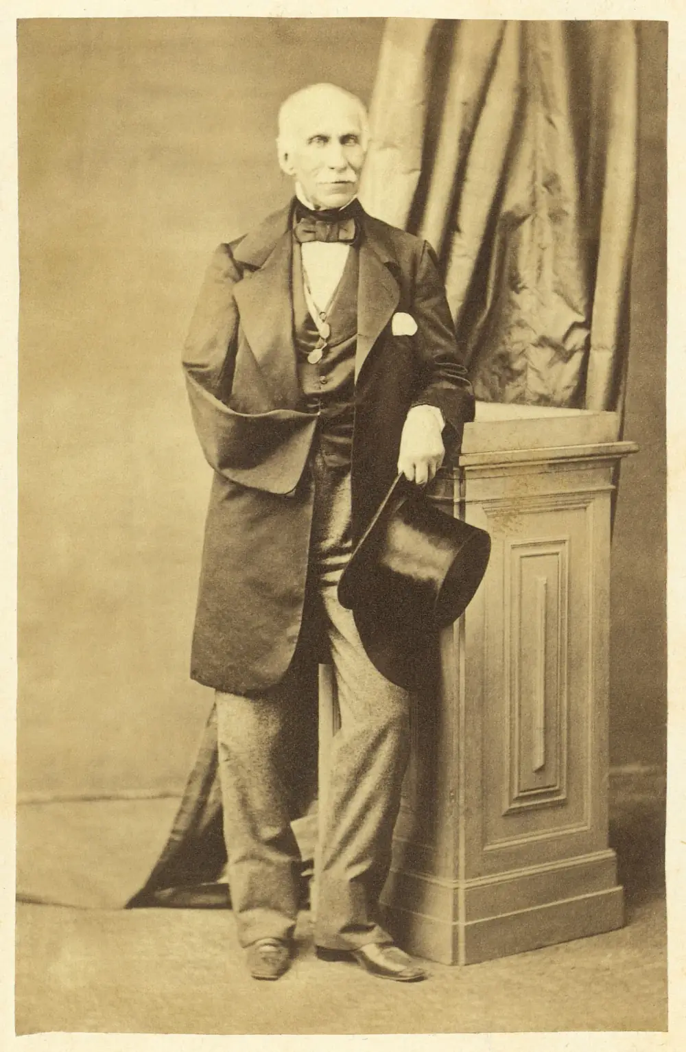 Sepia studio photograph of an elderly bald man with a white moustache, in a long dark coat and bow tie, standing beside a wooden pedestal and holding a top hat. His right sleeve hangs empty.