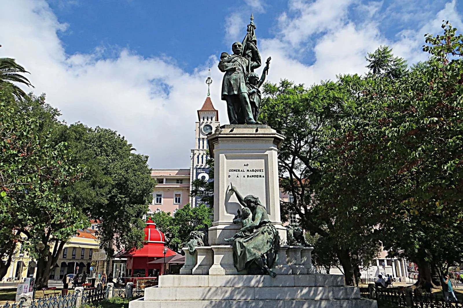 A bronze statue of a uniformed officer holding a flag, with a smaller figure at his side, stands on a tall stone plinth inscribed to General Marquis of Sá da Bandeira. At the foot of the plinth a seated bronze woman holding a child points up at him. Trees surround the square.