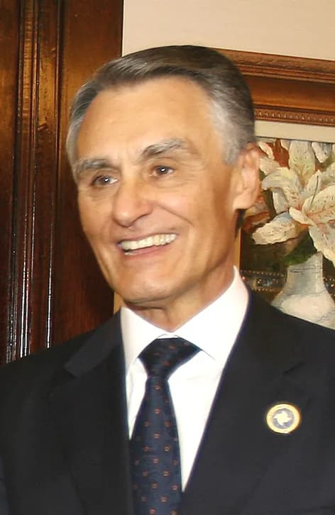 Head-and-shoulders portrait of a smiling grey-haired man in a dark suit, white shirt and patterned tie, in a wood-panelled room.