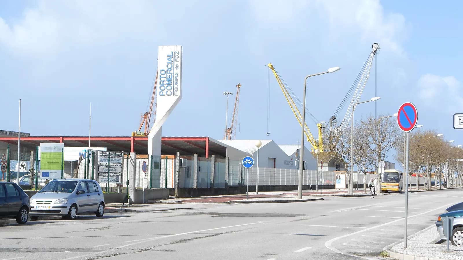 A tall white sign reading Porto Comercial Figueira da Foz stands at a fenced port entrance, with warehouses and yellow and white cranes behind and parked cars in the foreground.