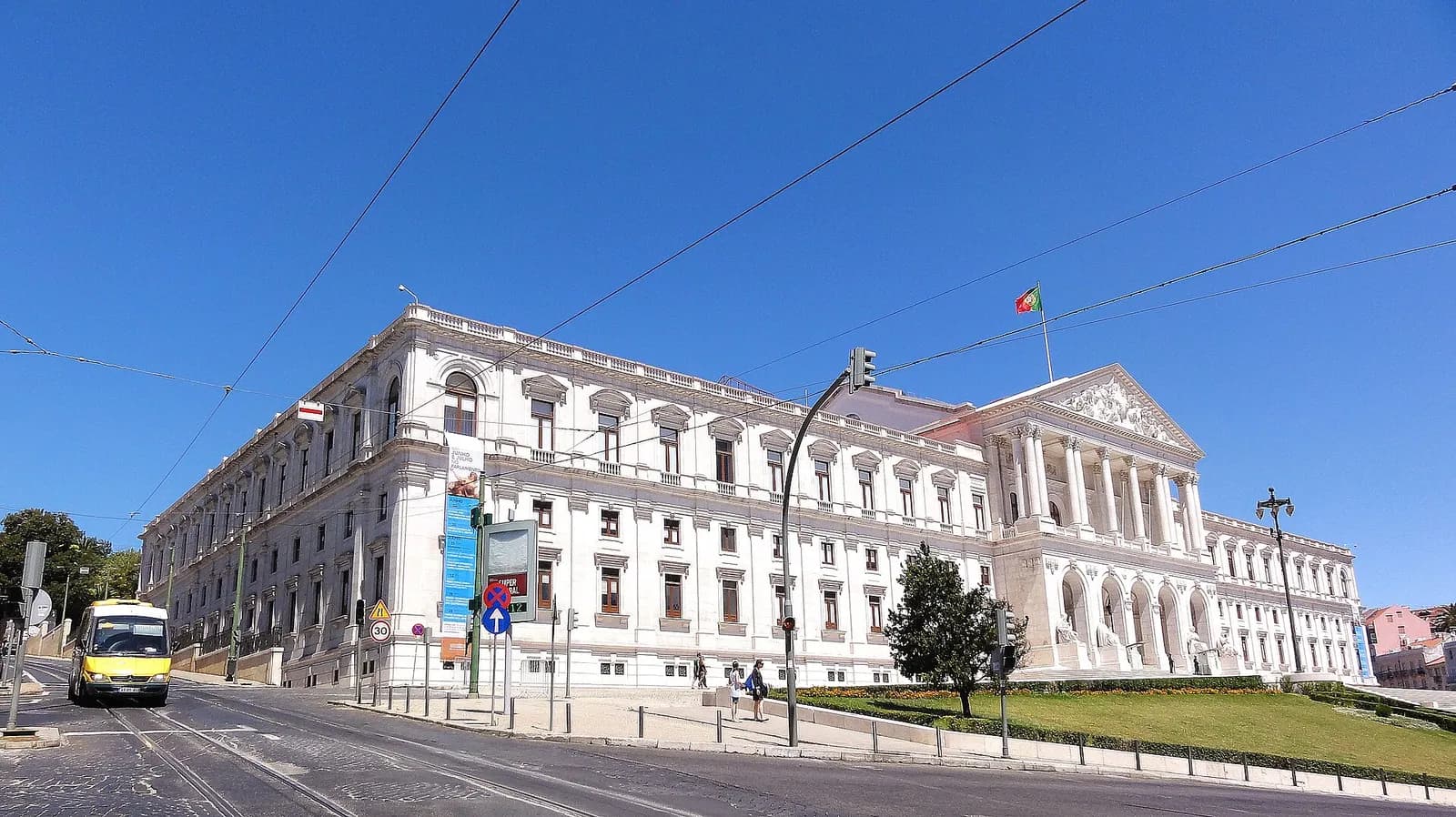 A long white neoclassical palace with a columned portico and a Portuguese flag on the roof, seen from a sunny street with tram wires overhead and a yellow minibus on the left.