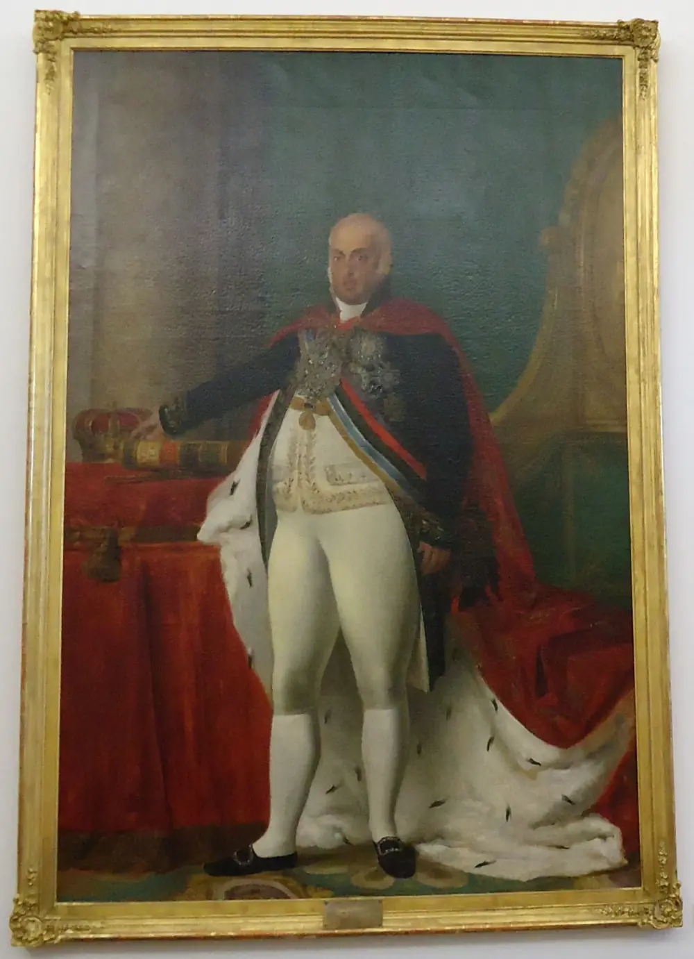 A full-length oil portrait in a gilt frame of a heavy-set, balding man in white breeches and stockings, a dark coat covered with orders and a red, ermine-lined royal mantle. His right hand rests on a book beside a crown on a red-draped table.