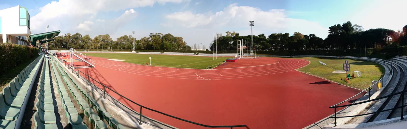 Panoramic view of an athletics stadium with a red running track around a green pitch, rows of empty green seats in the foreground and tall trees behind.