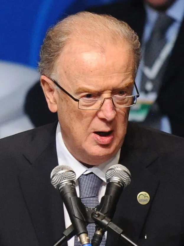 A grey-haired man in rimless glasses and a dark suit speaks into two microphones at a lectern.