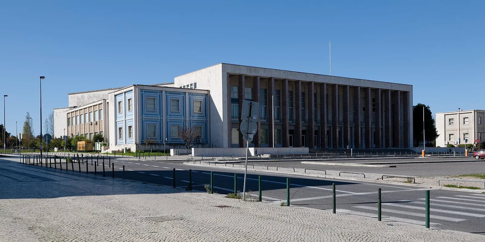 A long, flat-roofed white stone building with a portico of tall dark columns stands beyond an empty road and a cobbled pavement under a clear blue sky. A lower wing on the left is faced with blue and white tiles.