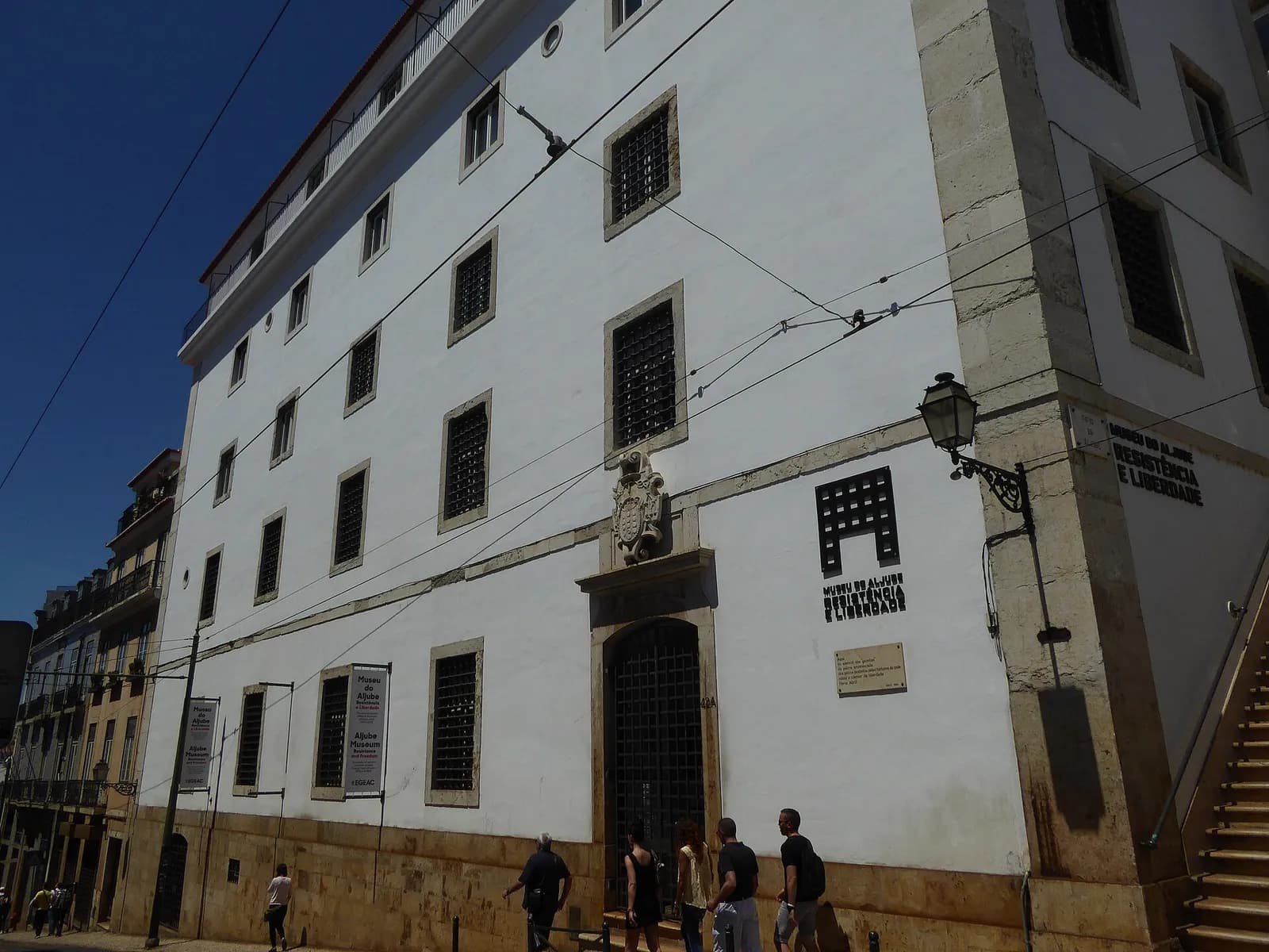 A tall whitewashed building with small barred windows and a stone coat of arms over an iron-grilled door, on a sunny Lisbon street with tram wires overhead and pedestrians passing.