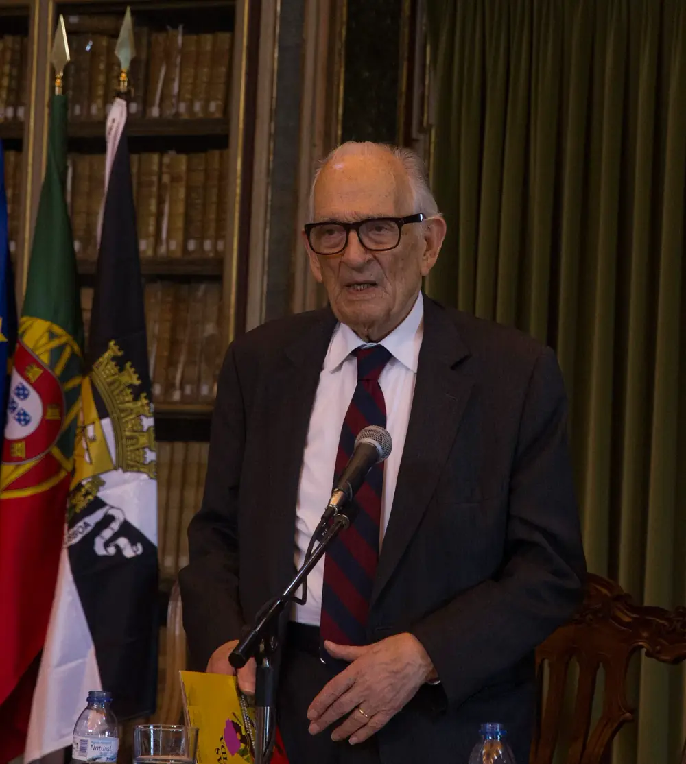 An elderly man with white hair and heavy black glasses, in a dark suit and striped tie, speaks into a microphone in front of bookshelves and the flags of Portugal, the European Union and Lisbon.