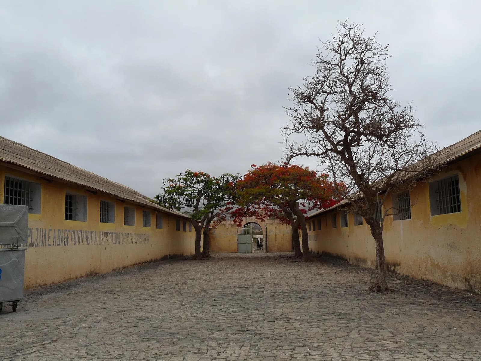 A cobbled yard runs between two long, low ochre buildings with barred windows towards an arched gateway with a metal gate, with a leafless tree, flame trees in red flower and a faded painted slogan on one wall, under a grey sky.