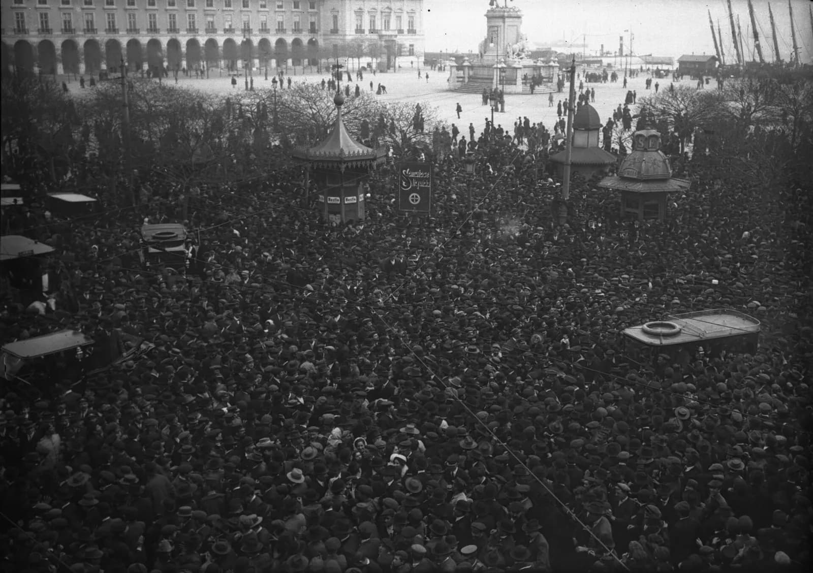 A dense crowd of men in hats packs a large square seen from above. Several motor cars are hemmed in by the crowd, and there are kiosks with pointed roofs among the people. In the background are arcaded buildings and an equestrian statue.