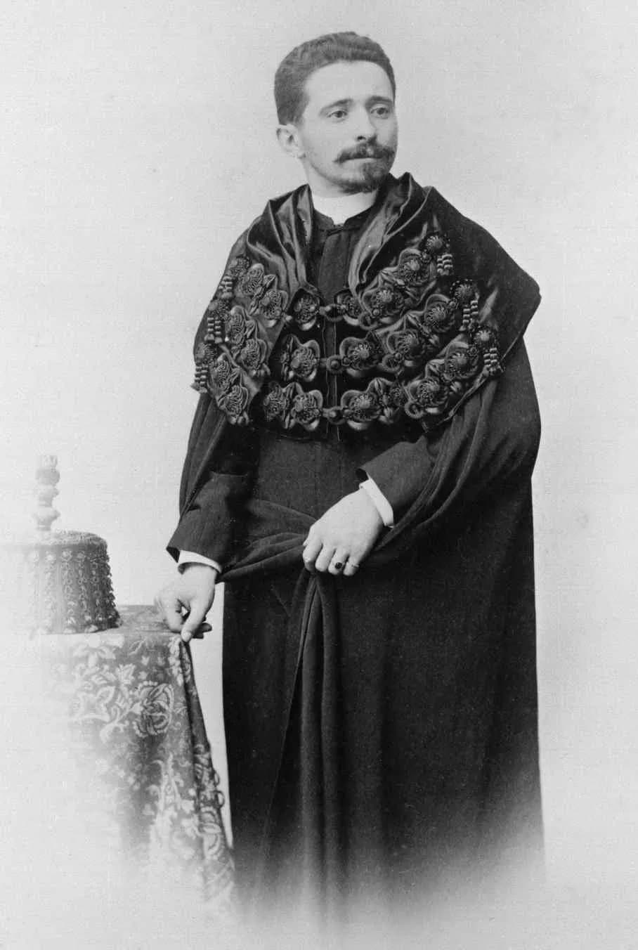 Studio portrait of a young man with short dark hair, a moustache and a small beard, standing beside a draped table on which rests a tasselled cap. He wears a long black gown and, over his shoulders, a short cape covered in rows of embroidered rosettes.