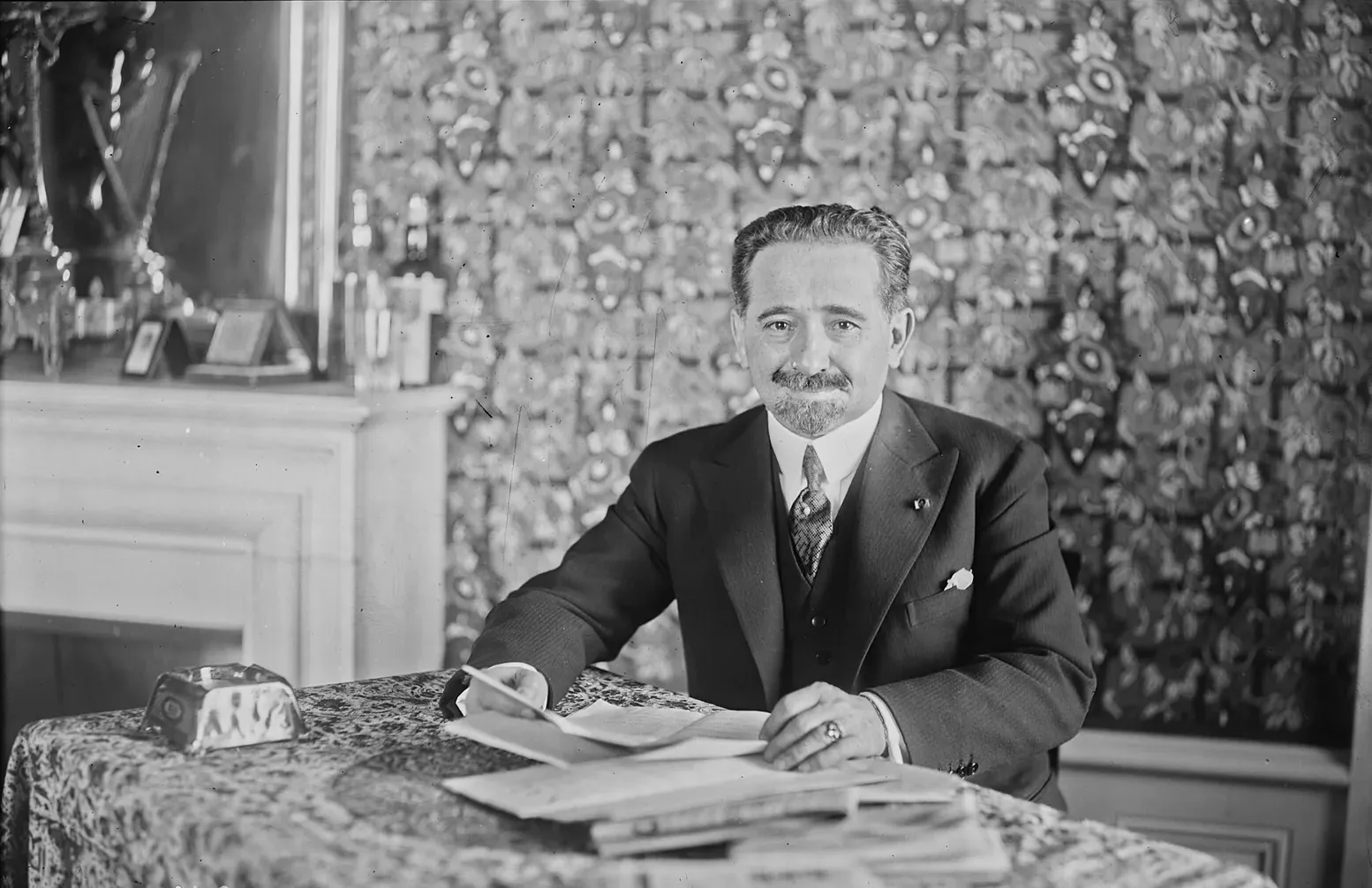 A smiling man with swept-back hair, a moustache and a small pointed beard sits at a table covered with a patterned cloth and loose papers, wearing a dark three-piece suit with a pin in the lapel. Behind him is a wall of patterned fabric and a white fireplace with bottles and framed pictures on the mantelpiece.